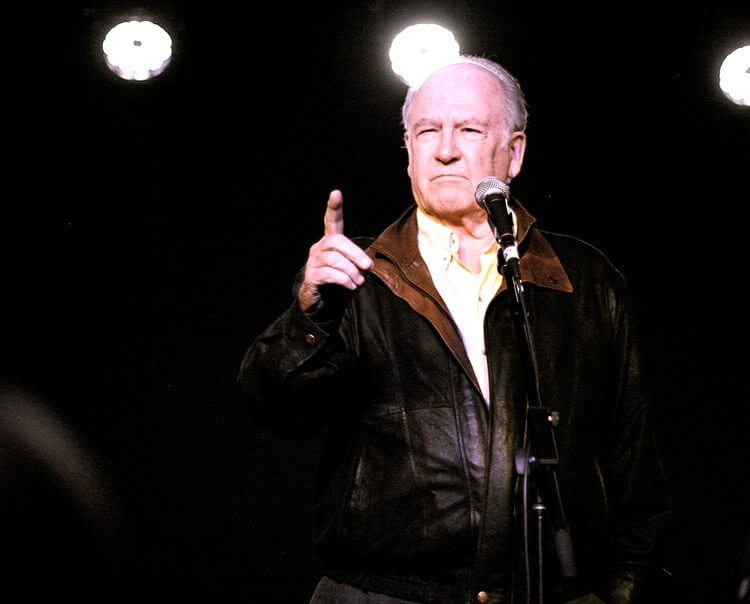 A man is standing in front of a microphone. The setting appears to be dark, suggesting an intimate atmosphere typical of a concert or gig. He is dressed in clothing suitable for an entertainment event, and his expression captures the focus of the moment.