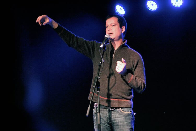 The image is of a man standing in front of a microphone, with three white lights above him against a dark background. He is wearing jeans and is standing while performing. The photo was taken by Amanda Peterson.