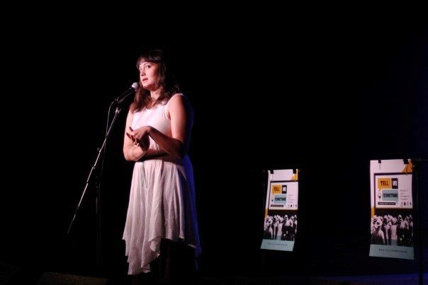 The image shows a person standing on a stage in front of a microphone, sharing a story. It is an indoor setting with stage equipment visible. There are two promotional posters displayed behind them. They are wearing a white dress. The image was taken by Amanda Peterson.