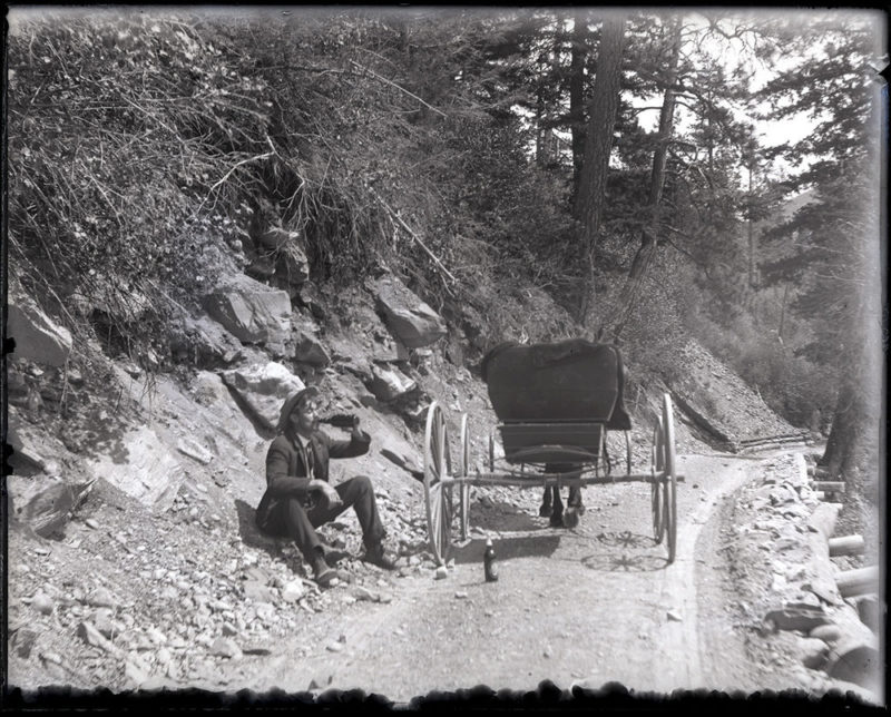 Man drinking beverage on side of dirt road. Horse drawn carriage next to him (via Montana Archives)