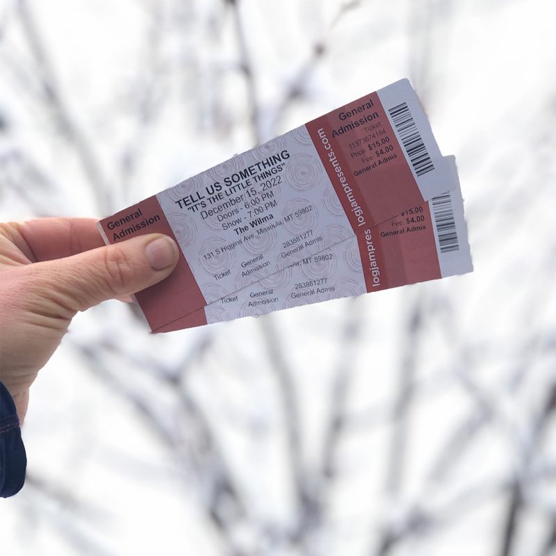a human holds two tickets to Tell Us Something "It's the Little Things" against a blurred wintery background