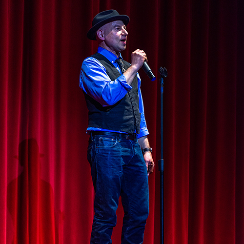 A man in a fedora stands in front of a red curtain with a microphone in his hand. photo credit Bob Ciampa