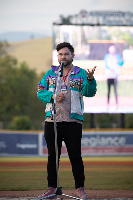 The photo shows a person standing on a stage holding a microphone and standing on grass. The background includes a clear sky and a depiction of the man on a large screen behind him. The image was taken by kmr studios.