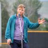 The image shows a person speaking into a microphone outdoors under the sky. They are standing and wearing blue trousers, a purple shirt and a teal jacket while speaking into a microphone. The photo was taken by KMR Studios.