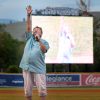 The image shows a person holding a microphone on a baseball field. The person is standing on the grass field and her image is also depicted on a screen behind her. The photo was taken by kmr studios.