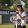 The image is of a person wearing glasses and gripping a microphone. The background shows outdoor scenery with trees and grass. The person is wearing athletic clothing and a hat. The photo was taken by kmr studios.