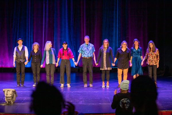 The image is a 9 people on a stage in a theater joining hands for a curtain call.