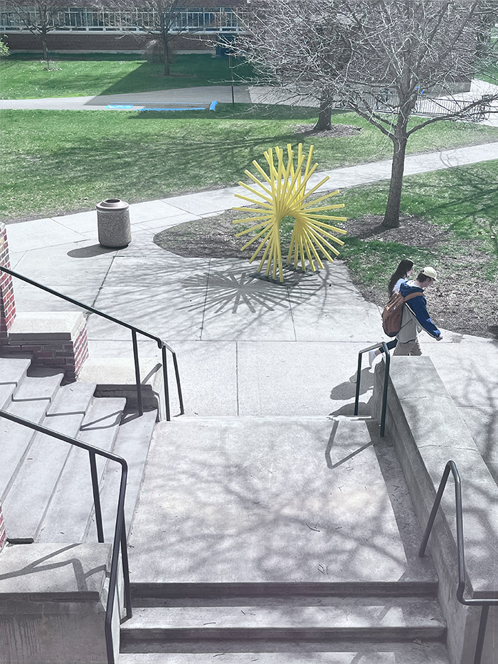 The image shows a couple of people walking on a sidewalk past a set of stairs on the campus of The University of Montana. There are trees and a bench in the background, as well as a spiky yellow metal sculpture resembling a dandelion flower.