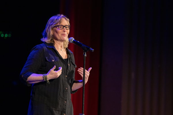 A woman with short blonde hair and glasses stands in front of a microphone sharing a story.