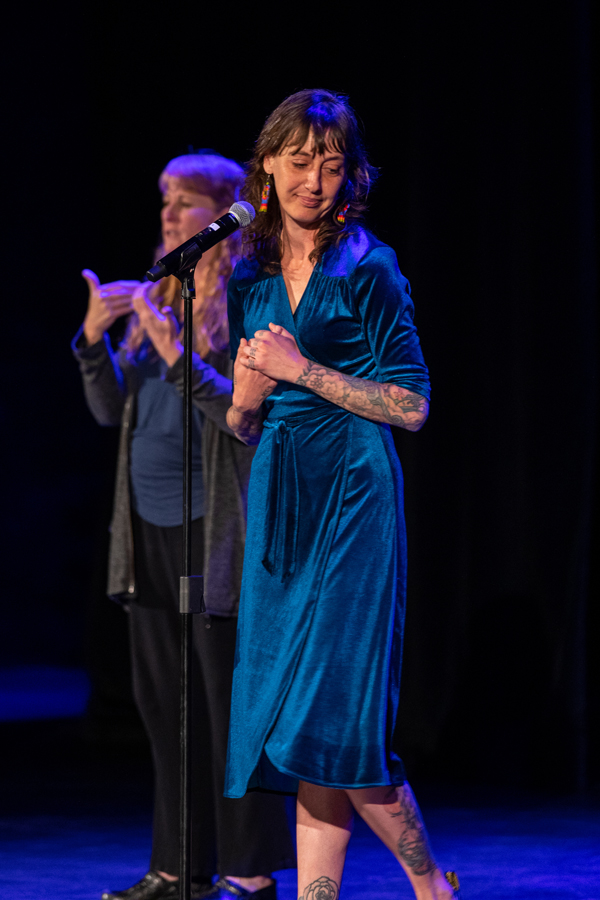 An ASL interpreter stands next to a woman in a blue dress. The woman in the dress has her head slightly turned away from a microphone as she arrives at an emotional part of the story she is sharing.