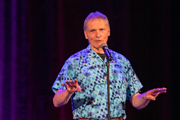 A man in a blue short sleeved shirt stands in front of a microphone sharing a story.