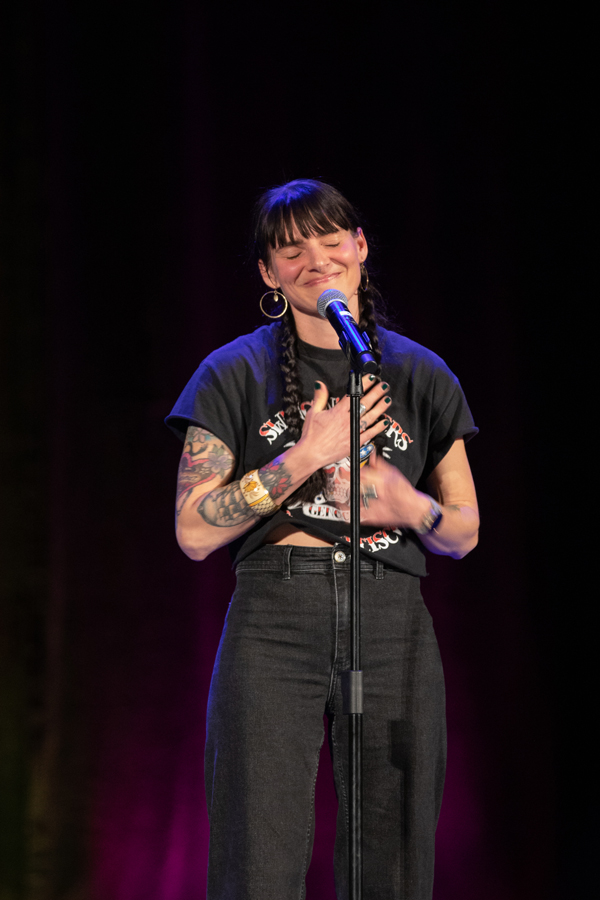 a woman with black hair cut into a style with bangs smiles as she places her hands opver her heart while she shares a story in front of a microphone.
