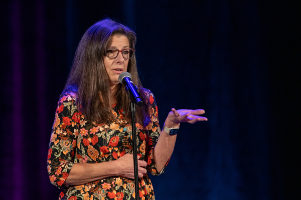 The image depicts a woman with long brown hair and glasses sharing a story in front of a microphone. The photo was taken by kmr studios.