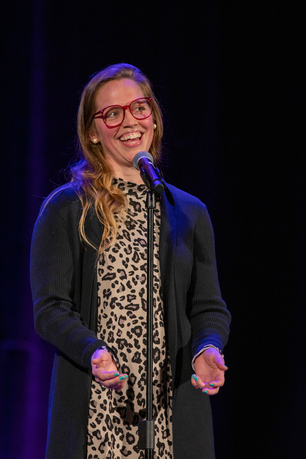The image depicts a woman with long sandy blonde hair and red glasses sharing a story in front of a microphone. The photo was taken by kmr studios.
