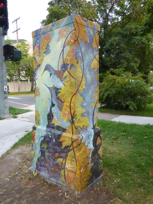 The image depicts a painting on a signal box outdoors. The painting shows a Flicker bird in a tree with grass and a blue sky in the background.