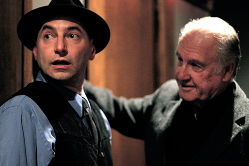 A man wearing a fedora hat is looking away from another man. The setting is indoors, and both men are dressed in fashionab...<p></p></div> <button class=