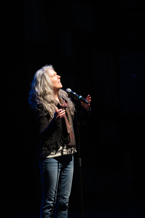 A woman with long hair is standing in front of a microphone, looking upwards. The image captures the energy of a live performance, showcasing her as an artist in the realm of entertainment. The photo is credited to KMR Studios.