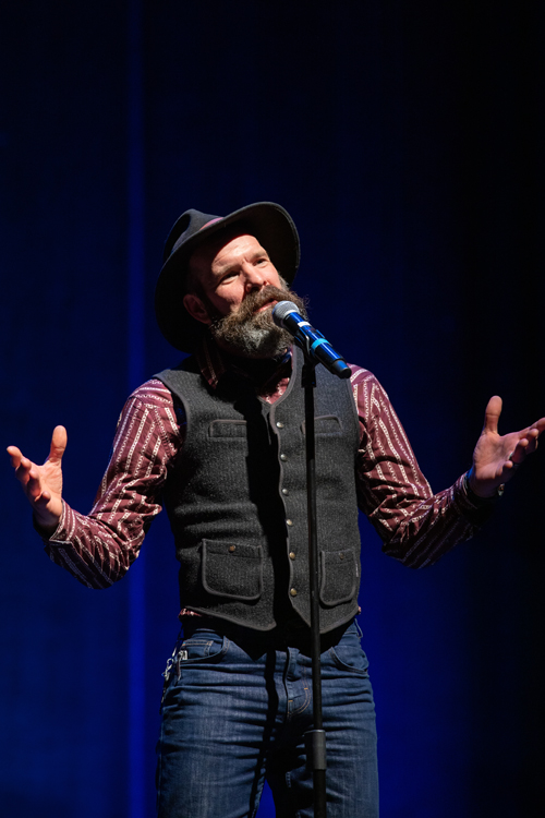 The image features a man with a beard wearing a hat, passionately speaking into a microphone. The scene captures the energy of live entertainment and the artistry involved in performance art. The photograph is taken by KMR Studios.