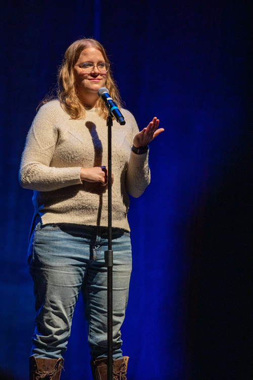 A woman is standing in front of a microphone. She is dressed in jeans and appears confident. The setting is indoors. The image was captured by kmr studios.