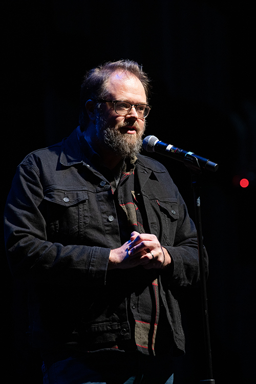 The image features a man with a beard speaking into a microphone. He is likely performing at an indoor storytelling event. The scene captures the energy of live entertainment and the artistry involved in performance art. The photograph is taken by kmr studios.