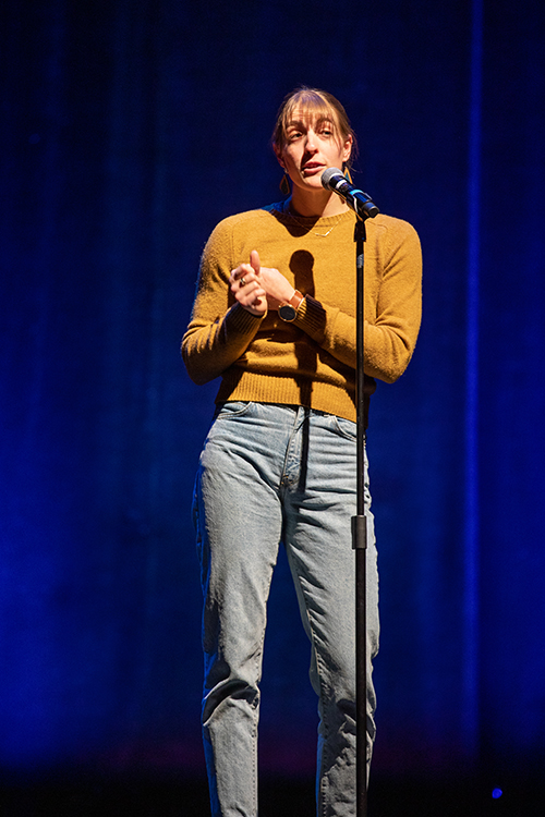 The image features a woman standing on a stage, holding a microphone. She seems to be engaged in a performance. The scene captures the essence of entertainment and storytelling, highlighting elements such as stage equipment and the art of telling a story. The photo by kmr studios.
