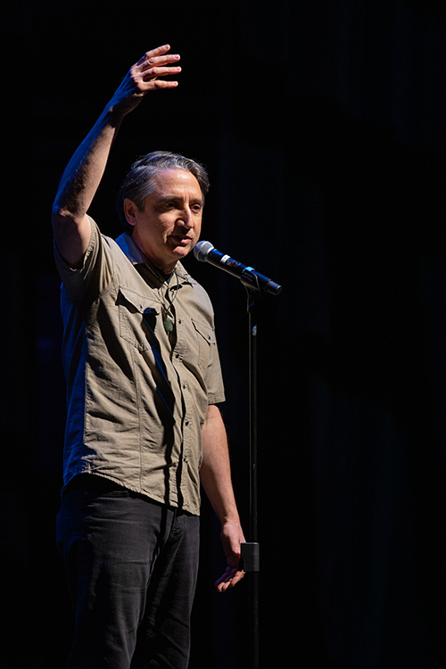 A man is standing in front of a microphone, likely at a concert or talent show. He appears to be addressing an audience. The setting includes audio equipment typical for live events. Photo by kmr studios.