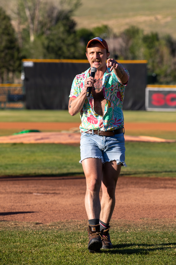 A man is holding a microphone outdoors, standing on grass. He is wearing a ballcap, a Hawaiian shirt, cutoff shorts and hiking boots. The scene is outdoors. Photo credit: kmr studios