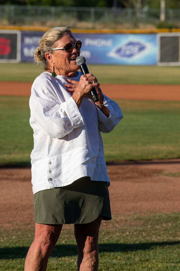 A woman is holding a microphone, sharing a story at Tell Us Something. The setting is outdoors. There are elements of grass and ground visible. The woman is dressed in a white shirt, green shorts and is wearing sunglasses. Photo credit: kmr studios