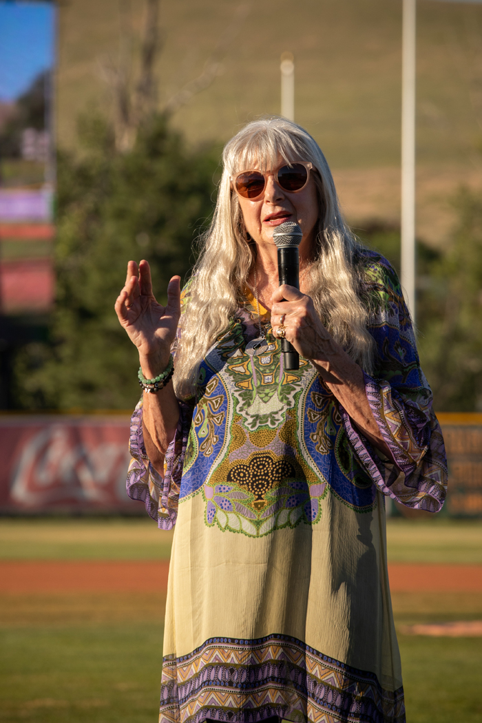 A woman is standing outdoors in a field, holding a microphone. She is wearing sunglasses and has her hair styled. The scene features green grass around her. she is wearing a patterned green dress. Photo credit: kmr studios
