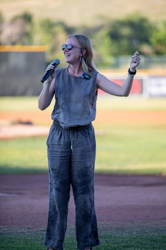 A woman is standing outdoors on a baseball field, holding a microphone. She is dressed in casual clothing wearing sunglasses and the setting features grassy areas. Photo credit: kmr studios