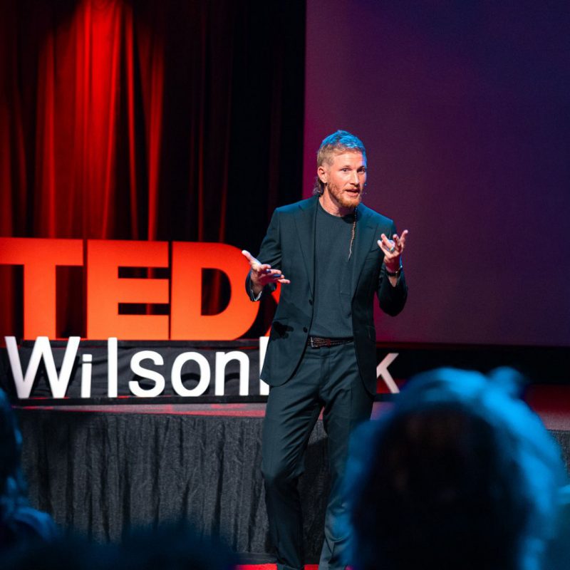 A man is standing on a stage, delivering a speech while a crowd watches attentively. He is dressed in a suit. The setting includes theater curtains and public address equipment.