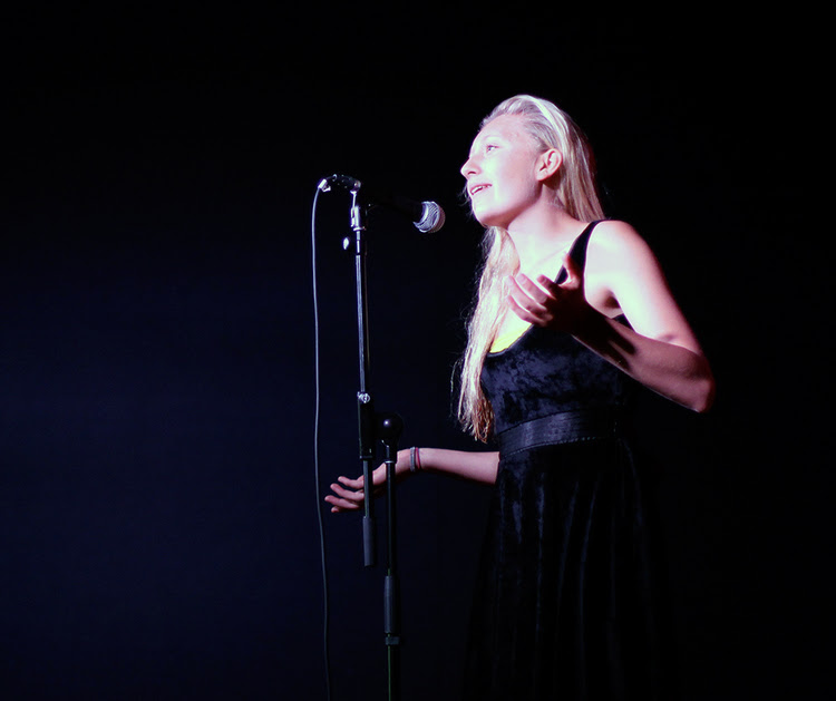 The image features a woman with long blonde hair sharing a story in front of a microphone. Photo credit - Amanda Peterson