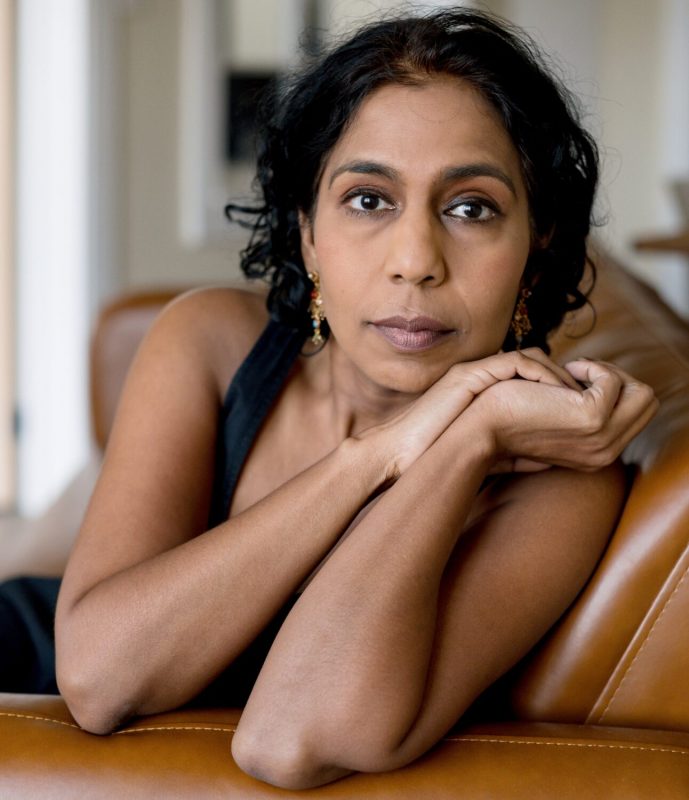 A woman with long hair is sitting on a couch indoors. She is dressed in fashionable clothing and poses as an art model for a photo shoot. The setting includes furniture and a wall in the background, highlighting her relaxed posture.