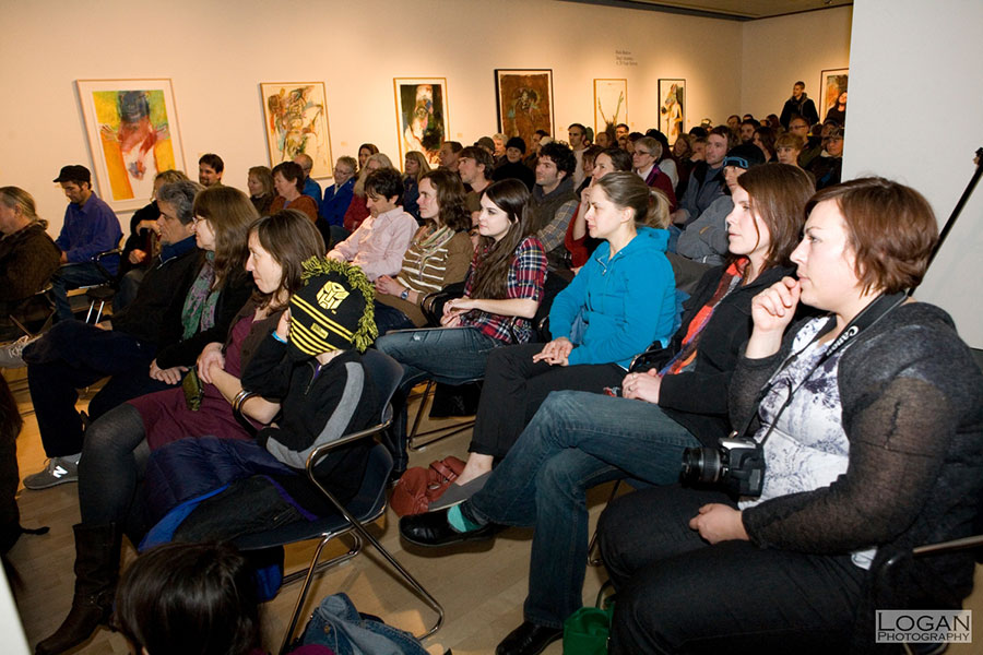A group of people is seated in chairs, arranged indoors. The scene includes individuals of various genders and clothing styles, listening to a storyteller who is out of the frame. The setting features a wall with art. Photo Credit Logan Photography