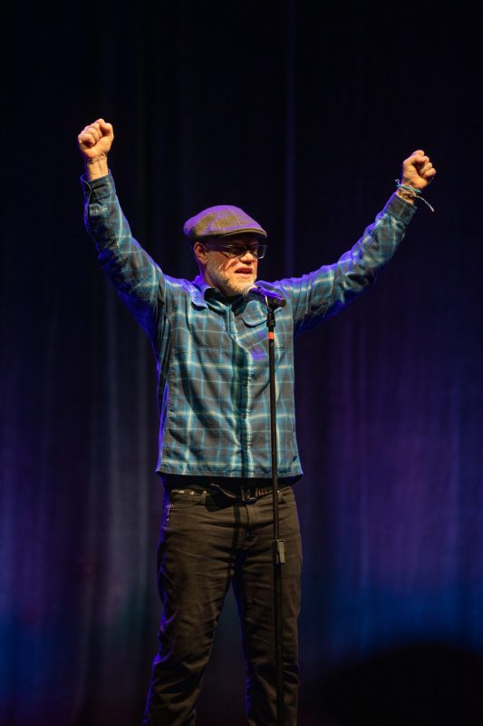 A person stands on stage with their arms raised in triumph. They are wearing a plaid shirt, a cap, and glasses. A microphone is positioned in front of them against a dark blue backdrop. 
