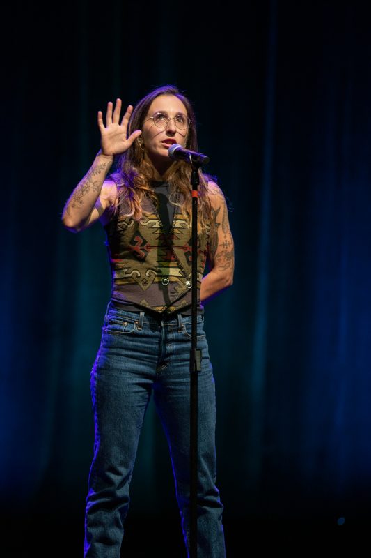 A person stands on stage with a microphone in front of them. They are wearing a sleeveless camouflage top and jeans. They have long hair and visible tattoos. The background is softly lit, creating a stage atmosphere. photo by kmr studios