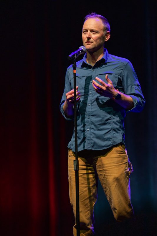 The image shows a man standing on stage in front of a microphone. He appears to be speaking or performing, with an expressive gesture using his hands. The background features soft lighting with red and blue hues, creating a dramatic atmosphere. The man is wearing a blue shirt and tan pants. photo by kmr studios