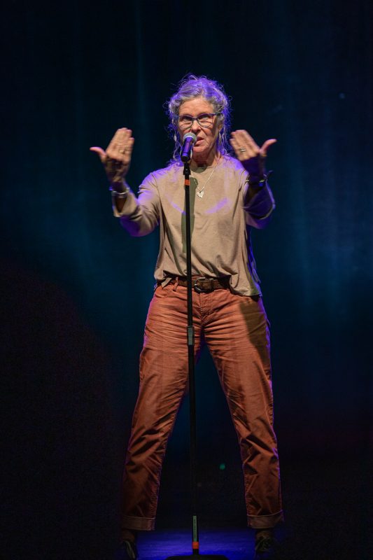 A person is performing on stage, standing in front of a microphone. They have medium-length hair and are wearing glasses, a light-colored top, and orange pants. The background features soft blue lighting that creates an engaging atmosphere for the performance. photo by kmr studios
